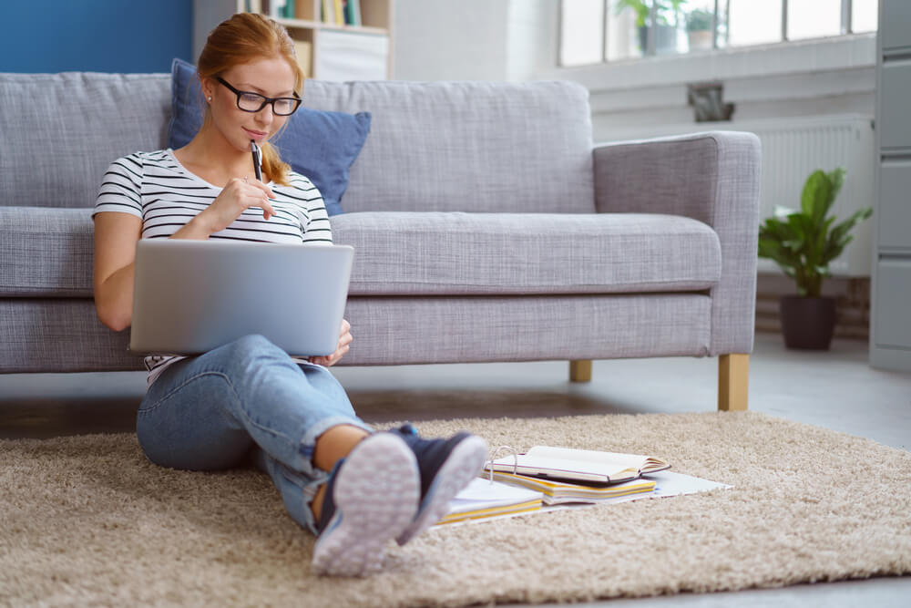 College student sits on floor of her apartment with back against the sofa and works on homework - cheap renters insurance for college students.